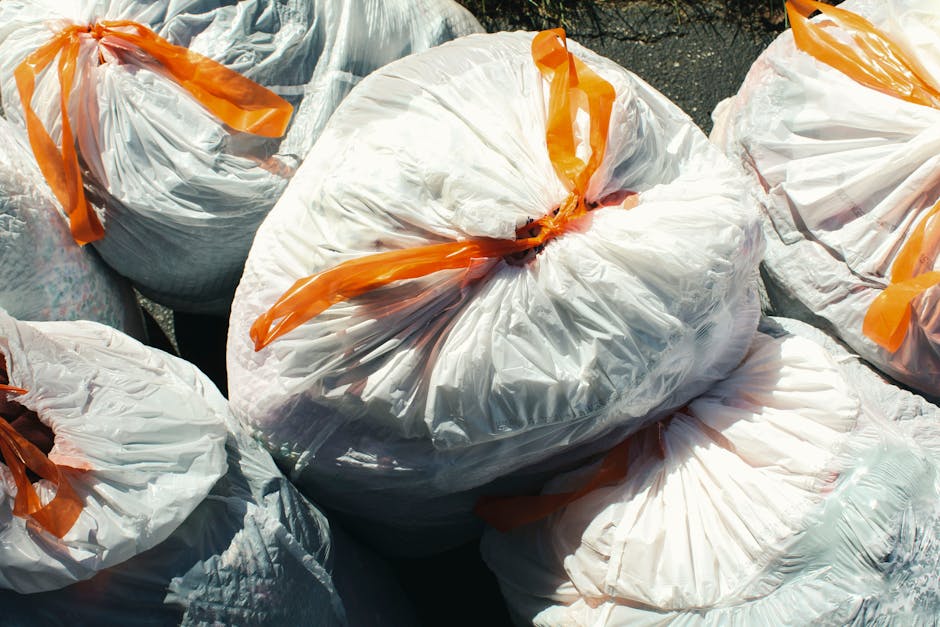 A person, wearing a brown jacket and dark shoes, is crouched on the ground outdoors, collecting litter. They are holding a clear plastic bottle with one gloved hand, about to place it into a brown pap